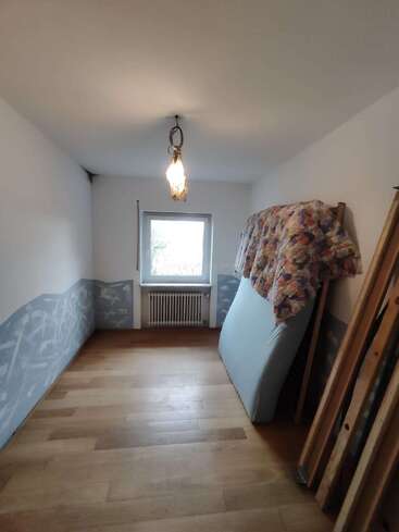 Una habitación pequeña y estrecha con suelo de madera, una sola ventana y una lámpara de techo. Un colchón y la ropa de cama están apoyados contra la pared, junto a las piezas desmontadas de una cama de madera.