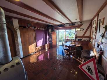 Comedor luminoso con vigas de madera, suelo de baldosas brillantes, decoración vintage, mesa redonda, asientos acogedores, reloj de pie, grandes ventanas con vistas al jardín y una decoración ecléctica en las paredes.