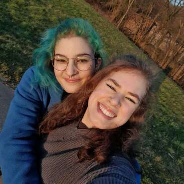 Two friends pose for a cheerful selfie outdoors. One has blue-green hair and glasses, the other is smiling brightly. Sunlight gently illuminates their happy faces.