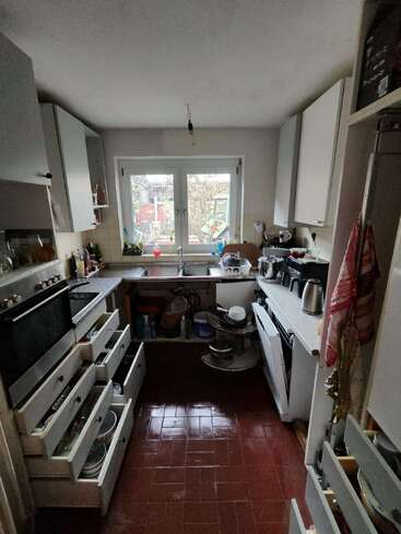 Esta cocina tiene suelo de baldosas marrones, armarios blancos y cajones en su mayoría abiertos, una ventana grande, un fregadero, electrodomésticos de encimera, vajilla y utensilios esparcidos por todas partes, dando una sensación de desorden.