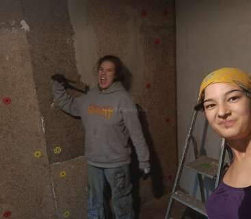 Dos mujeres están reformando una habitación. Una de ellas sonríe para hacerse un selfi, con un pañuelo amarillo en la cabeza, mientras que la otra trabaja con entusiasmo en la pared con una herramienta.