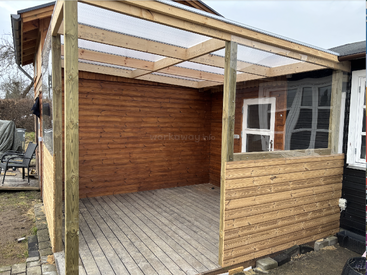 This image shows a wooden patio structure attached to a house. The patio has a clear plastic roof, wooden floor, and is partially enclosed with timber panels.