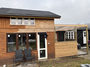 This image shows a small wooden house with large windows, two black chairs outside, and an attached partially-constructed covered patio area on the right.