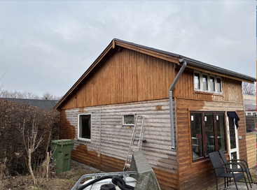 The image shows a wooden house being stained; the upper parts are a rich brown while the lower areas are still gray, with a ladder leaning nearby.