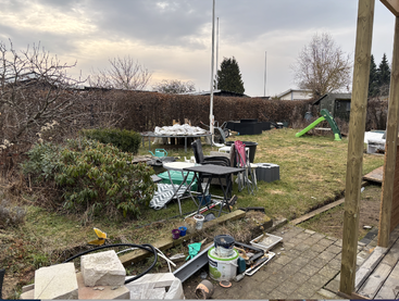 A cluttered backyard with construction materials, paint cans, scattered chairs, a table, garden tools, a green slide, bushes, and overcast sky, suggesting ongoing renovations.