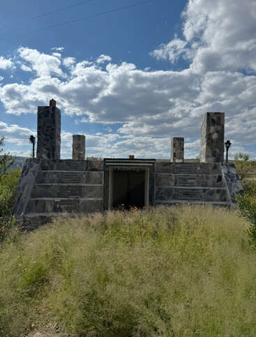 A stone building partially embedded in the ground with four tall pillars, surrounded by wild grass under a sky filled with scattered, fluffy clouds.