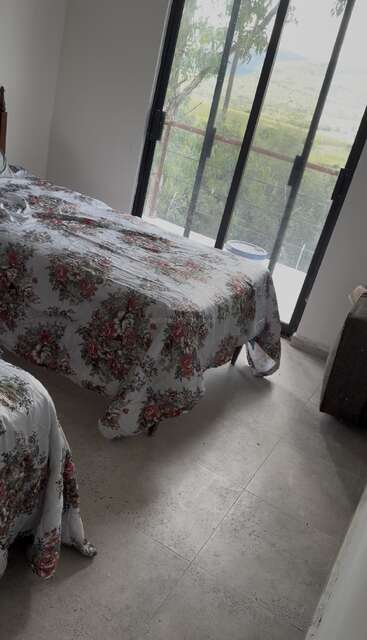 A bright bedroom with two beds featuring floral bedspreads, tile flooring, large glass doors, and a scenic outdoor view of greenery and hills beyond. Peaceful atmosphere.