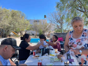 A family enjoys an outdoor meal by a pool under bright blue skies. Children and adults sit together, sharing food and drinks on a sunny day.