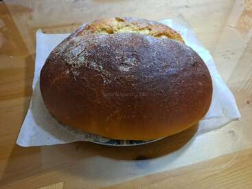 Um pão redondo, de cor marrom-dourada, está sobre uma folha de papel manteiga em cima de uma mesa de madeira. A crosta está levemente rachada e polvilhada com farinha, parecendo recém-assado.