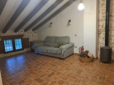 Esse aconchegante quarto no sótão tem um sofá cinza, vigas de madeira rústicas no teto, paredes de pedra, piso de ladrilhos de barro, fogão a lenha, uma cesta de lenha e uma janela pequena.