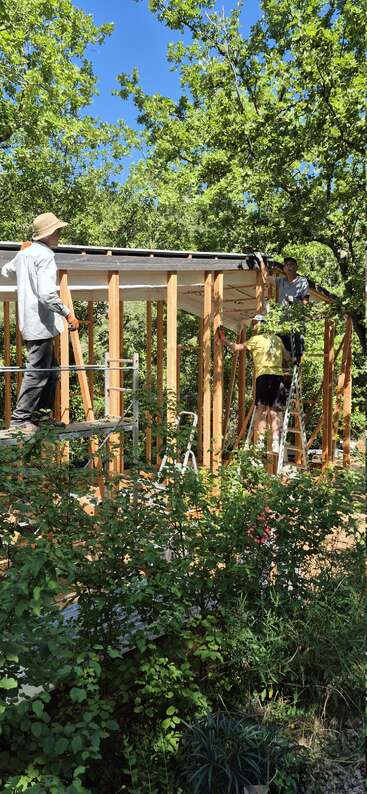 Three people are building a wooden structure in a lush, green forest. Sunlight filters through the trees as they work on the framing and roof.