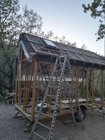 A small wooden house or tiny home is under construction outdoors, with exposed framing, roof work in progress, a window, and two ladders leaning against it.