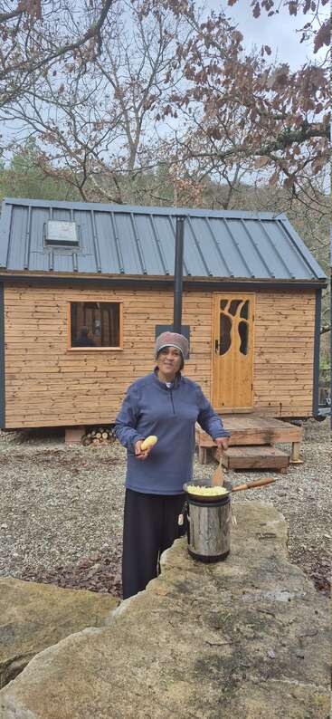A woman in a blue jacket and beanie is cooking outdoors, preparing food on a portable stove, with a wooden tiny house and leafless trees behind her.