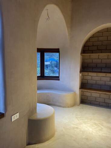 Esta imagen muestra una acogedora sala minimalista con paredes curvas de adobe, bancos empotrados, nichos arqueados, estanterías de ladrillo y una ventana con vistas al jardín.