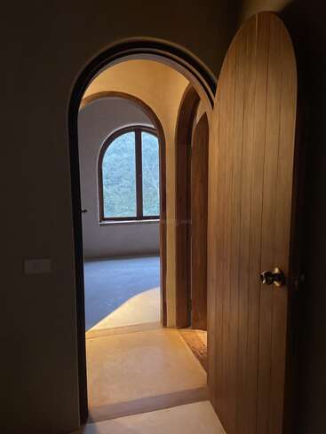 Una puerta de madera arqueada, cálidamente iluminada, se abre a un pasillo, revelando otra puerta arqueada y una ventana con vistas a una exuberante vegetación, creando un ambiente sereno y acogedor.