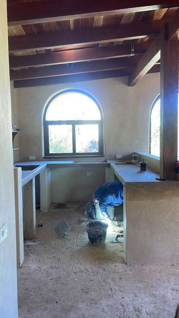 Un trabajador vestido de azul se arrodilla bajo una encimera en una cocina rústica e inacabada con ventanas arqueadas, vigas de madera y herramientas de construcción esparcidas por el suelo.