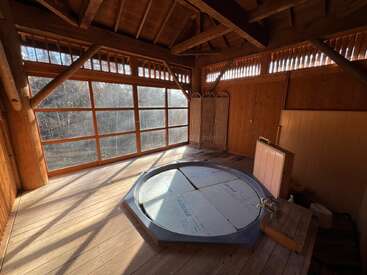 Esta imagen muestra una habitación de madera iluminada por el sol, con grandes ventanales, una bañera de hidromasaje redonda con cubierta, techos altos y un biombo decorativo en la esquina.