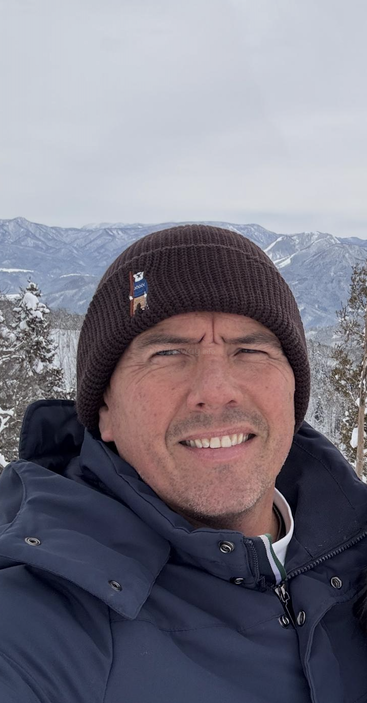 Un hombre con una chaqueta oscura de invierno y un gorro marrón se toma una selfie al aire libre. Árboles nevados y montañas lejanas forman un fondo invernal.