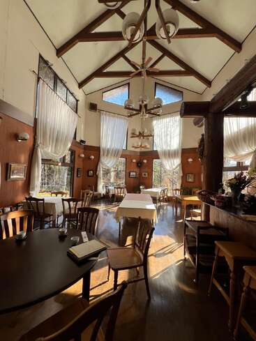 La luz del sol se cuela por grandes ventanales con cortinas de visillo en un restaurante rústico y acogedor. Vigas de madera, lámparas de araña y mesas dispersas crean un ambiente cálido y acogedor.