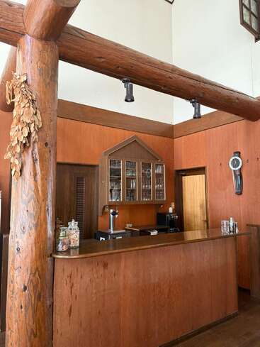 Este acogedor bar de madera cuenta con vigas de troncos rústicos, vitrinas, una cafetera, botes de aperitivos, hierbas secas y una cálida iluminación natural, que evocan un ambiente acogedor.