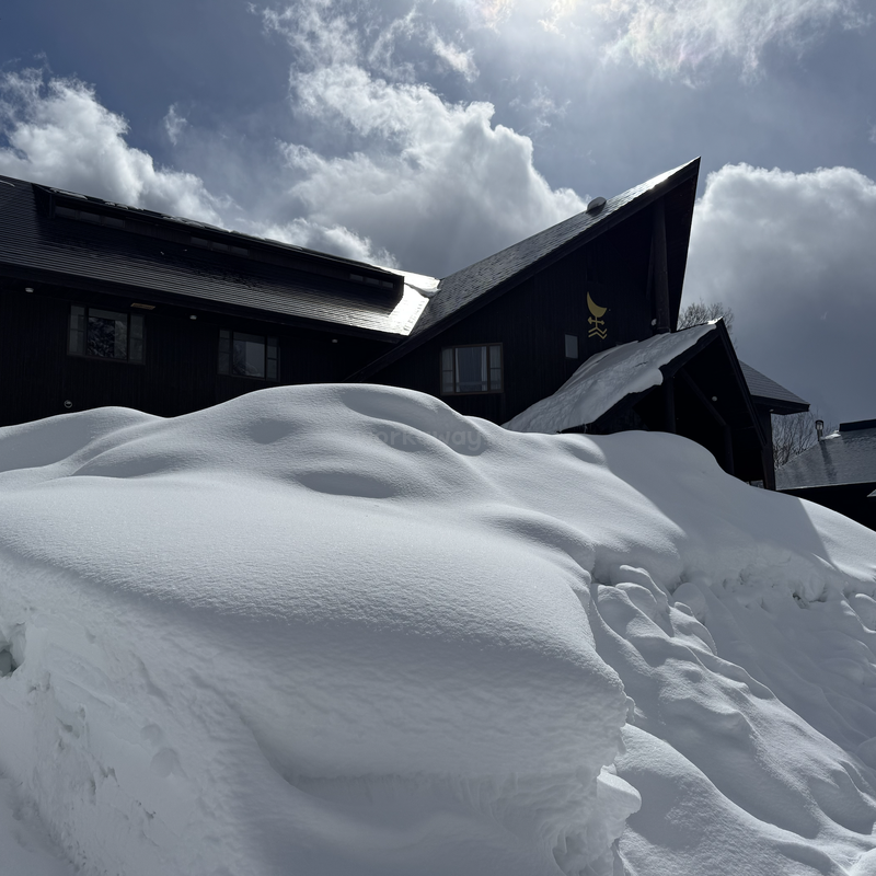 Un edificio negro con tejados empinados se encuentra detrás de un gran montículo de nieve fresca e intacta bajo un espectacular cielo nublado en el que se filtra la luz del sol.
