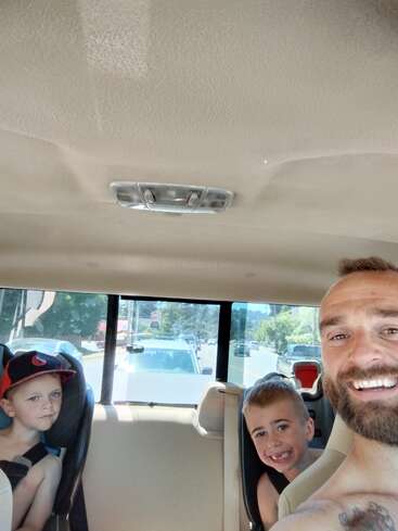 Un homme et deux jeunes garçons sont à l'intérieur d'une voiture, souriant et profitant de leur temps ensemble. Les garçons sont assis dans des sièges de voiture ; la lumière du soleil brille à l'intérieur.