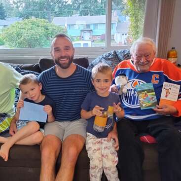Une photo de famille joyeuse montre trois générations : deux enfants, leur père et leur grand-père, qui porte un maillot de hockey. Ils tiennent des cartes, des livres et une peluche.