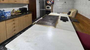 Una cocina con armarios de madera, encimera de mármol, plátanos, botellas de agua, fogones, microondas, frigorífico, sofá, TV en la pared, banco y una tableta.