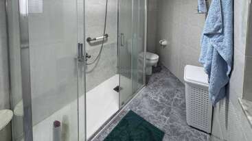 Esta imagen muestra un cuarto de baño moderno con cabina de ducha de cristal, inodoro montado en la pared, toallas azules, cesto de la ropa sucia, alfombrilla verde y suelo y paredes de baldosas.