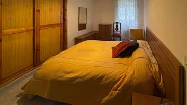 Este dormitorio cuenta con una cama grande con funda amarilla, muebles de madera, un armario, un espejo, una cómoda, una silla individual y una ventana con cortinas.