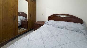 Esta imagen muestra un acogedor dormitorio con una cama de madera, una colcha gris claro, una mesilla de noche de madera y un gran armario con puertas de espejo que reflejan los muebles.