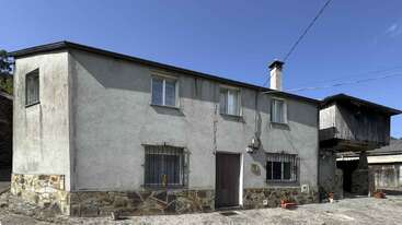 Esta imagen muestra una casa de dos plantas con cimientos de piedra, ventanas enrejadas y una extensión superior de madera. El exterior aparece erosionado, bajo un cielo azul despejado.