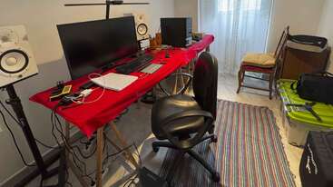 Una oficina en casa con un gran monitor, teclado, altavoces y varios aparatos electrónicos sobre un escritorio cubierto de rojo. Hay sillas, contenedores y una alfombra de rayas.