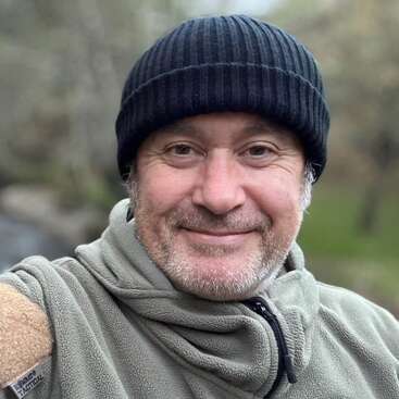 Un homme au sourire amical porte un bonnet noir et une veste polaire verte. Il se tient à l'extérieur, avec un arrière-plan naturel flou derrière lui.