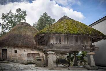 Esta imagen muestra dos antiguos edificios rústicos con gruesos tejados de paja cubiertos de musgo y zócalos de piedra, rodeados de árboles, bajo un cielo azul nublado.