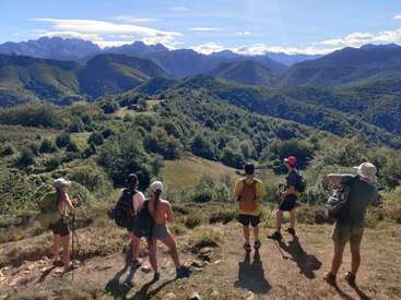 Sechs Wanderer mit Rucksäcken und Mützen stehen auf einem grasbewachsenen Hügel und genießen den Panoramablick auf die üppig grünen Berge unter einem strahlend blauen Himmel.