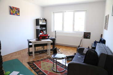 A bright room features a keyboard, guitar, bookshelf, sofa, small tables, rug, microphone stand, backpack, and abstract art, creating a cozy music practice space.