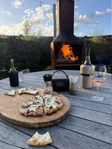 Una deliciosa pizza sobre una tabla de madera descansa sobre una mesa rústica al aire libre, rodeada de bebidas, condimentos y un fuego crepitante al fondo.