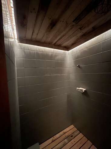 This image shows a minimalist shower with dark tiles, a wooden ceiling, and a wooden floor. There's subtle lighting along the ceiling edges, creating a cozy atmosphere.
