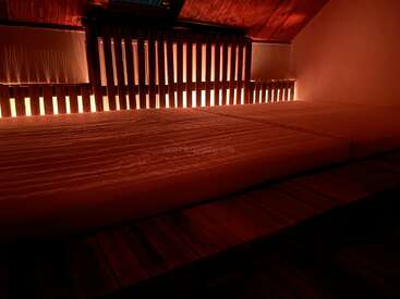 A cozy loft bedroom with a large mattress on a wooden platform, softly illuminated by warm, ambient lighting from behind wooden slats along the wall.