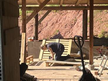 A person kneels on the ground working on a wooden pallet under a roofed structure. Sunlight streams in, highlighting tools, wood, and construction materials scattered around.