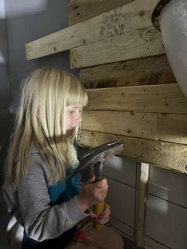 A young girl with blonde hair holds a hammer, working intently on a wooden wall. Light and shadows emphasize her concentration and the texture of the wood.