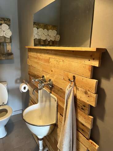 This rustic bathroom features a wooden accent wall, vintage sink, large mirror, rolled towels on shelves, toilet, and a white towel hanging beside the sink. Cozy atmosphere.