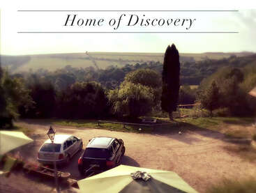 A serene countryside landscape with cars parked on a gravel area, green trees, rolling hills, and soft sunlight. Text above reads "Home of Discovery."