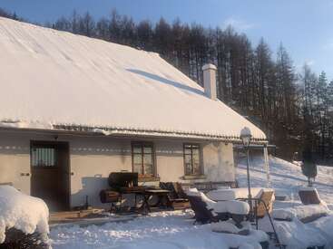 A charming snow-covered house stands in a peaceful winter landscape. Outdoor furniture and a grill are blanketed with snow. Pine trees line the background, bathed in sunlight.