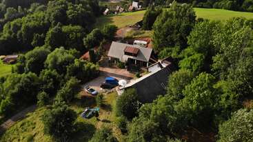 This aerial photo shows a countryside house surrounded by dense green trees, vehicles parked outside, a small inflatable pool, and open grassy fields in the background.