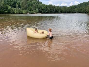 Three children play in a shallow lake. Two sit inside a yellow inflatable raft while the third pushes it. Forested hills and trees surround the tranquil water.