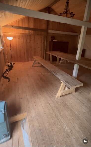 Cette image montre une pièce rustique en bois avec de longs bancs et des tables, un lampadaire, des murs en bois, des poutres apparentes et un lustre suspendu au plafond.
