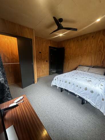 Este quarto aconchegante tem paredes com painéis de madeira, uma cama grande com edredom estampado, carpete cinza, ventilador de teto, TV, armário aberto e duas portas abertas.