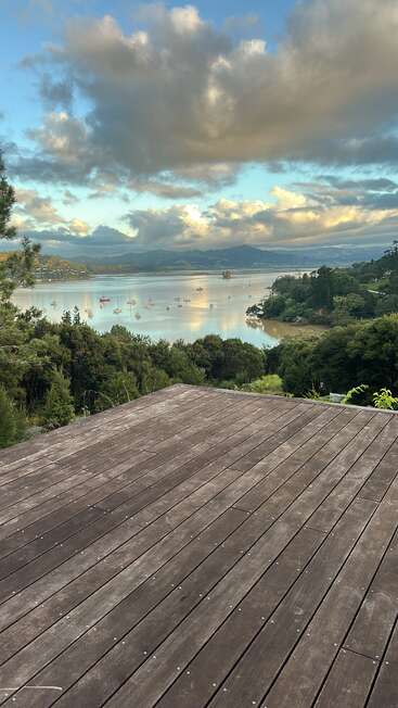 Um deck de madeira tem vista para uma baía tranquila com veleiros, cercada por colinas verdes e árvores, sob um céu noturno nublado e espetacular, com a luz suave do sol.